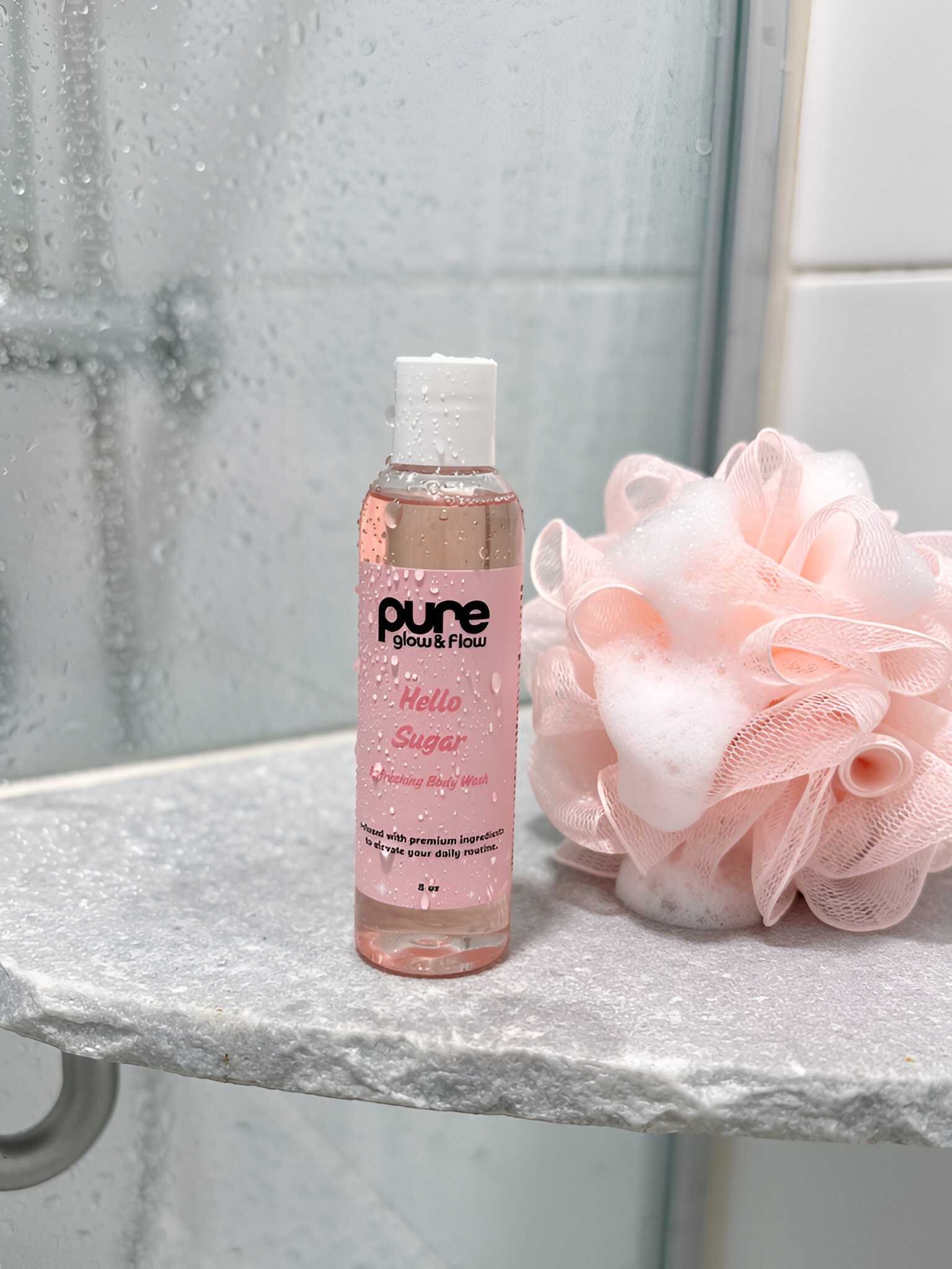 Bottle of 'pure glow & flow Hello Sugar' scrub with a pink bath pouf on a bathroom shelf.